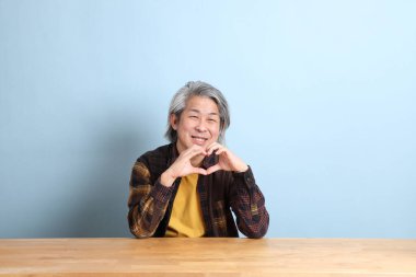 The senior Asian man wearing the yellow plaid shirt sitting at the working desk with blue background.