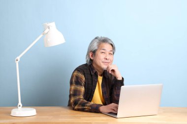The senior Asian man wearing yellow plate shirt working on laptop at the working desk with the blue background.