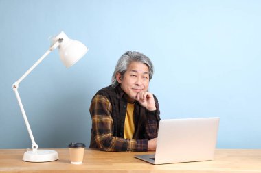The senior Asian man wearing yellow plate shirt working on laptop at the working desk with the blue background.