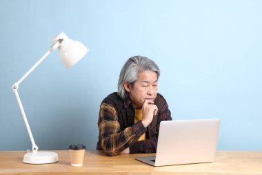 The senior Asian man wearing yellow plate shirt working on laptop at the working desk with the blue background.