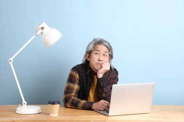 The senior Asian man wearing yellow plate shirt working on laptop at the working desk with the blue background.