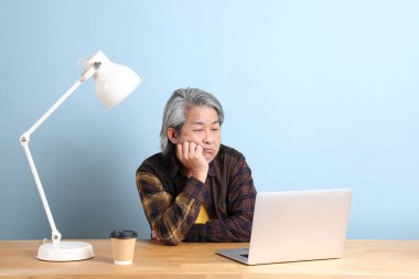 The senior Asian man wearing yellow plate shirt working on laptop at the working desk with the blue background.