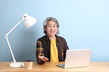 The senior Asian man wearing yellow plate shirt working on laptop at the working desk with the blue background.