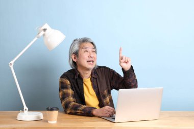 The senior Asian man wearing yellow plate shirt working on laptop at the working desk with the blue background.