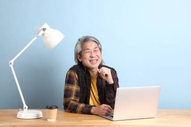 The senior Asian man wearing yellow plate shirt working on laptop at the working desk with the blue background.