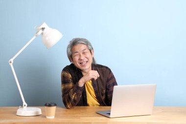The senior Asian man wearing yellow plate shirt working on laptop at the working desk with the blue background.