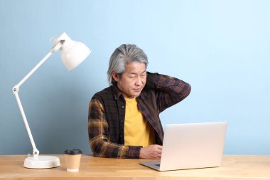 The senior Asian man wearing yellow plate shirt working on laptop at the working desk with the blue background.