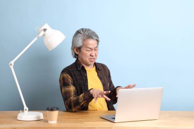 The senior Asian man wearing yellow plate shirt working on laptop at the working desk with the blue background.