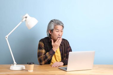 The senior Asian man wearing yellow plate shirt working on laptop at the working desk with the blue background.