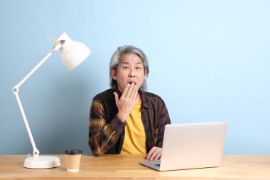 The senior Asian man wearing yellow plate shirt working on laptop at the working desk with the blue background.