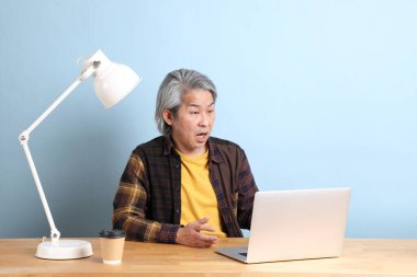 The senior Asian man wearing yellow plate shirt working on laptop at the working desk with the blue background.