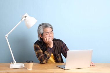 The senior Asian man wearing yellow plate shirt working on laptop at the working desk with the blue background.