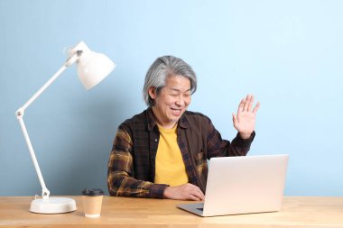 The senior Asian man wearing yellow plate shirt working on laptop at the working desk with the blue background.