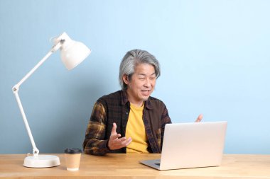 The senior Asian man wearing yellow plate shirt working on laptop at the working desk with the blue background.