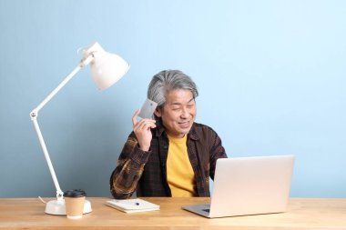 The senior Asian man wearing yellow plate shirt working on laptop at the working desk with the blue background.