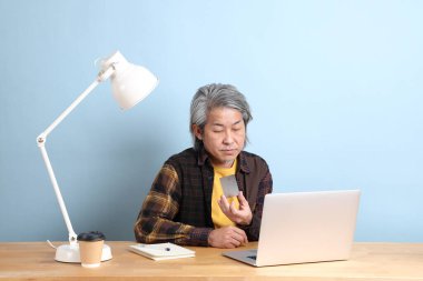 The senior Asian man wearing yellow plate shirt working on laptop at the working desk with the blue background.