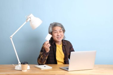 The senior Asian man wearing yellow plate shirt working on laptop at the working desk with the blue background.