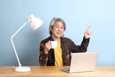 The senior Asian man wearing yellow plate shirt working on laptop at the working desk with the blue background.