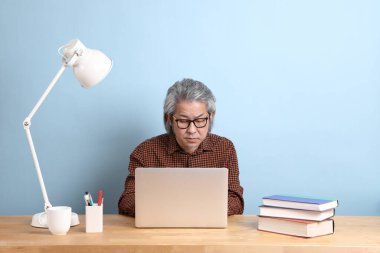 The senior Asian man working on the laptop at the desk with blue background.