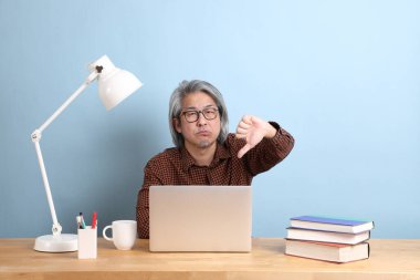 The senior Asian man working on the laptop at the desk with blue background.