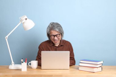 The senior Asian man working on the laptop at the desk with blue background.