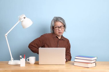 The senior Asian man working on the laptop at the desk with blue background.