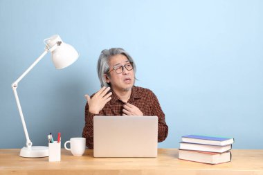 The senior Asian man working on the laptop at the desk with blue background.