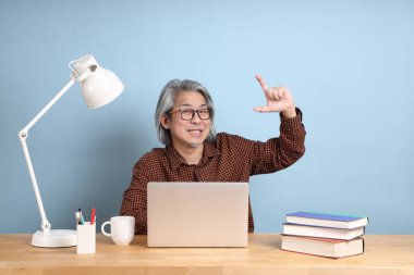 The senior Asian man working on the laptop at the desk with blue background.