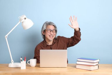 The senior Asian man working on the laptop at the desk with blue background.