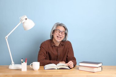 The senior Asian man reading the books at the desk with the blue background.