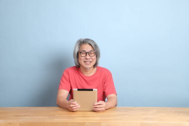 The senior Asian man with colorful shirt sitting at the desk with blue background.