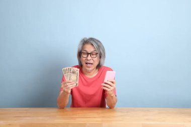 The senior Asian man with colorful shirt sitting at the desk with blue background.