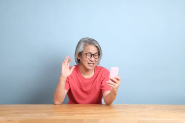 The senior Asian man with colorful shirt sitting at the desk with blue background.