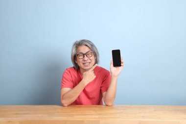 The senior Asian man with colorful shirt sitting at the desk with blue background.
