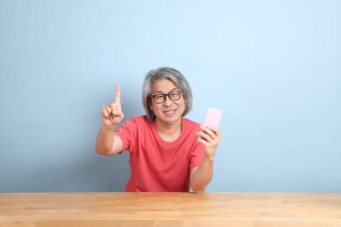 The senior Asian man with colorful shirt sitting at the desk with blue background.