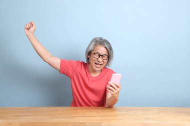 The senior Asian man with colorful shirt sitting at the desk with blue background.