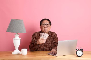 The senior Asian man sitting at working desk with laptop on the pink background.