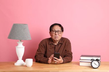 The senior Asian man sitting at working desk using smart phone on the pink background.