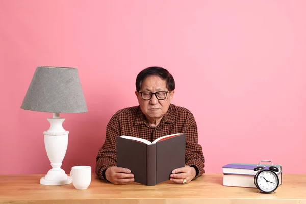 Senior Asian Woman Reading Book Sitting Desk Pink Background — Stockfoto