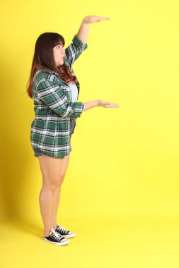 Sarı arka planda sıradan kıyafetlerle duran tombul Asyalı kadın..