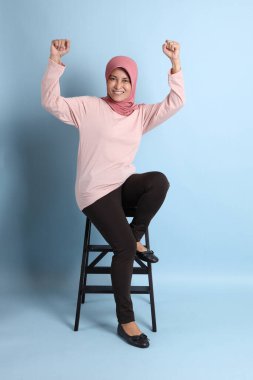 Mavi arka planda oturan tesettürlü Güneydoğu Asyalı yaşlı kadın.