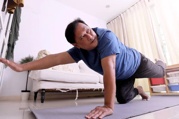 40多岁的亚洲成年超重男子在电视上看体育节目 并在客厅的地图上做瑜伽 — 图库照片