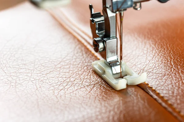 Modern sewing machine presser foot with a needle sews brown leather. Sewing process of decorative seam on leatherette with special leather presser foot. Close up, copy space.