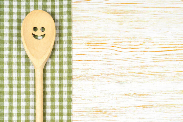 Wooden spoon with smile on green checkered tablecloth on white wooden surface. Mockup for menu or recipe, restaurant, website with cooking. Kitchen food background, template, flat lay with copy space