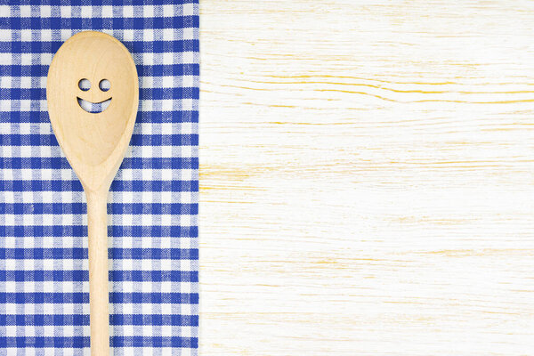 Деревянная ложка с улыбкой на голубой клетчатой скатерти на белой деревянной поверхности. Макет для меню или рецепт, ресторан, веб-сайт с кулинарией. Пищевой фон Китчен, соблазнительный, плоский, с пространством для копирования
