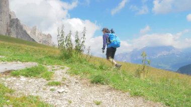Belluno, Ağustos 2020. Tam teçhizatlı kadın yürüyüşçü güneşli bir günde yüksek dağdaki bir patikanın yakınındaki yeşil çimlerde yürüyor. Arka plandaki dağlarda mavi gökyüzünde