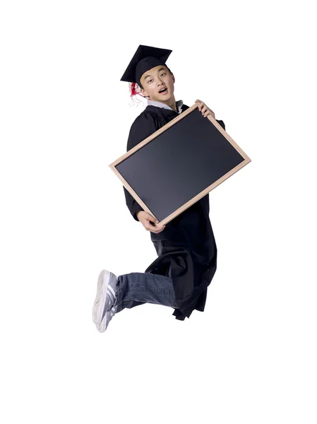 Male graduate jumping while holding a black board — Stock Photo, Image