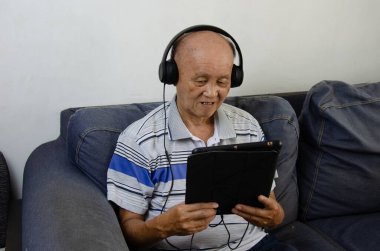 Kanepede elinde tablet olan yaşlı bir adam kulaklık ve sosyal medya ile müzik dinliyor..