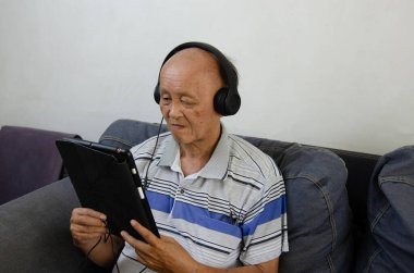 Kanepede elinde tablet olan yaşlı bir adam kulaklık ve sosyal medya ile müzik dinliyor..
