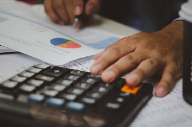 Elinde hesap makineleri ve iş belgeleri bulunan işadamı istatistiksel veri yatırım analizi ve vergi muhasebesi grafikleri tutuyor.